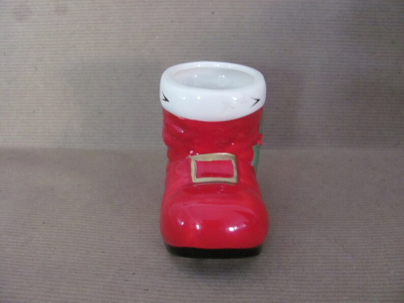 May include: A ceramic Santa boot-shaped container. The boot is primarily red with a white cuff and a gold-colored buckle. Green accents are visible on the side. The container has a black base and is designed for decorative or functional use.