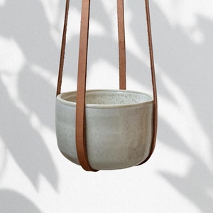May include: A white ceramic hanging planter with a light brown leather strap. The planter is hanging from the ceiling.