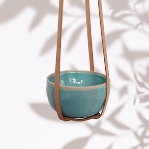 Può includere: Un vaso sospeso con una ciotola in ceramica turchese e cinghie in pelle marrone chiaro. La ciotola ha un bordo color beige chiaro ed è sospesa a tre cinghie in pelle. Lo sfondo è bianco con ombre di foglie.