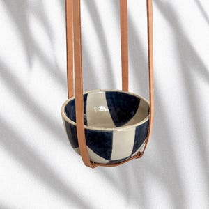 Könnte beinhalten: Eine hängende Keramikschale mit blau-weißem geometrischem Design. Die Schale wird von hellbraunen Lederriemen gehalten. Der Hintergrund ist eine weiße Wand mit Schatten.
