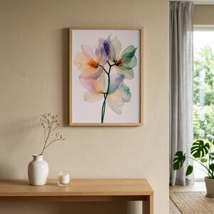 May include: A framed floral print featuring translucent flowers in shades of peach, blue, and green. The artwork is displayed on a beige wall above a wooden table. A white vase with a sprig and a candle are on the table.