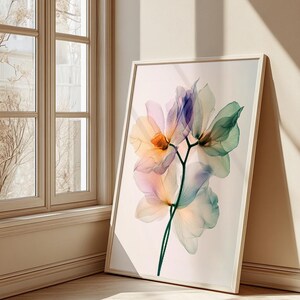 May include: A framed floral art print featuring a delicate, translucent flower with petals in shades of orange, pink, purple, and green. The artwork is set against a neutral background and leans against a wall near a window.