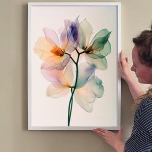 May include: A framed floral art print featuring a translucent flower design. The artwork showcases petals in shades of orange, purple, and green, set against a cream background. The print is encased in a white frame, suitable for home decor.