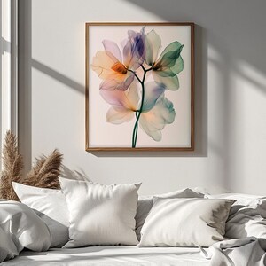 May include: A framed floral art print featuring delicate, translucent flowers in shades of orange, purple, and green. The artwork is set against a light background and framed in a natural wood frame. The print is displayed above a bed with white pillows and bedding.