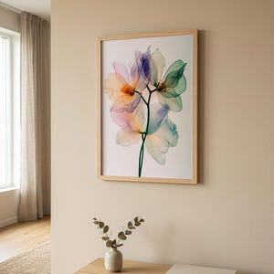 May include: A framed floral art print featuring translucent flowers in shades of purple, orange, green, and blue. The artwork is displayed in a light wood frame and hangs on a beige wall. A small vase with greenery sits on a wooden cabinet below.