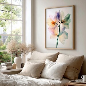 May include: A framed floral art print featuring translucent flowers in shades of pink, yellow, purple, and green. The artwork hangs above a bed with beige pillows and white bedding. A window with a view of greenery is visible.