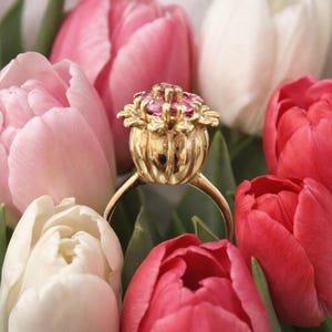 May include: A gold ring with a floral design, featuring a cluster of pink gemstones at the centre. The ring is set against a backdrop of pink, red, and white tulips, creating a vibrant and elegant composition. The ring's intricate details are highlighted.