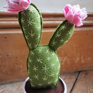 May include: A felt cactus with two pink flowers in a white pot. The cactus is green with white embroidered details.