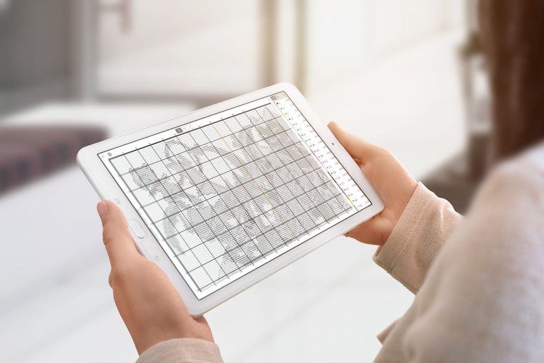May include: A person is using a tablet to view a cross-stitch pattern. The pattern is displayed on the screen in black and white, with a grid of squares. The tablet is white and has a black bezel.