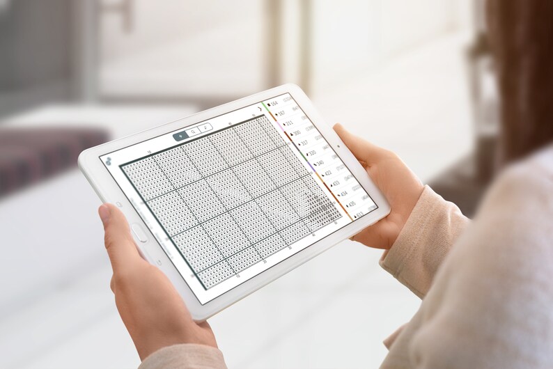 May include: A white tablet displaying a cross-stitch pattern on the screen. The pattern is a grid of small squares, with some squares filled in with different colours. The tablet is being held by a person's hands.