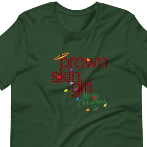 Puede incluir: Una camiseta verde oscuro con las palabras "brown skin girl" en letras rojas a cuadros y un halo dorado. Las palabras "Merry Christmas" están en verde con luces navideñas coloridas.