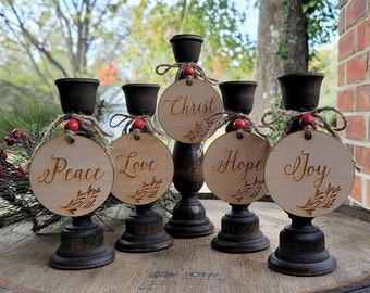May include: Five wooden candle holders with a rustic finish, each with a round wooden tag tied with twine and a red bead. The tags are inscribed with the words "Peace", "Love", "Christ", "Hope", and "Joy".