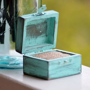 May include: A teal blue wooden ring box with a hinged lid and a burlap lining. The box is distressed and has a rustic look.