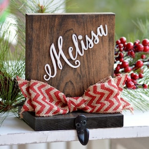 May include: A wooden Christmas stocking holder with the name "Melissa" written in white paint. The holder has a red and white chevron patterned burlap bow and a black metal hook.