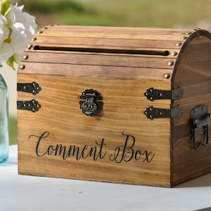 May include: A wooden comment box with a curved top and decorative metal hardware. The box is made of light brown wood and features the words "Comment Box" in elegant script. A slot is cut into the top for inserting cards.