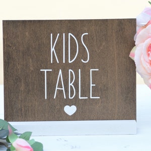 May include: A wooden sign with the words "KIDS TABLE" in white lettering and a white heart symbol. The sign is brown with a white base and is likely for a children's event or area.