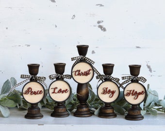 May include: Five wooden candle holders with black and white checkered ribbon and round wooden tags with the words "Christ", "Peace", "Love", "Joy", and "Hope" in red lettering.