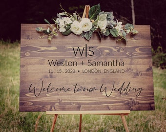 May include: A rustic wooden wedding sign with white flowers on top. The sign reads "W|S Weston + Samantha 11.15.2023 • LONDON ENGLAND Welcome to our Wedding"