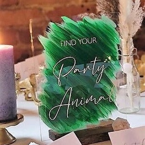 May include: A green acrylic sign with white brushstrokes and the text "Find Your Party Animal" in white cursive font. The sign is on a wooden stand and there are two gold dinosaur figurines in the foreground.