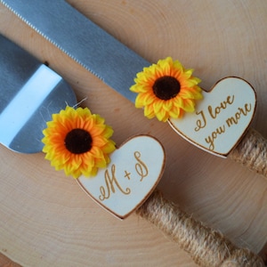 May include: A set of two cake cutting knives with wooden handles wrapped in twine. Each handle is decorated with a sunflower and a heart-shaped wooden plaque. One plaque says "I love you more" and the other says "M + S".