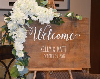 May include: A wooden welcome sign with white lettering, decorated with a garland of white flowers and greenery. The sign reads "Welcome", "Kelly & Matt", and "October 21, 2017".