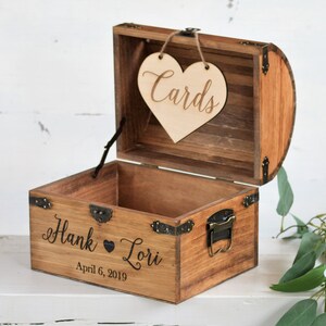 Puede incluir: Un cofre de madera con un letrero en forma de coraz&oacute;n que dice "Cards" colgando de la parte superior. El cofre est&aacute; grabado con los nombres "Hank & Lori" y la fecha "April 6, 2019".