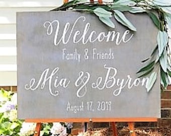 May include: A gray wooden sign with white lettering that reads "Welcome Family & Friends Mia & Byron August 17, 2019". The sign is decorated with green leaves and is propped up on an easel.
