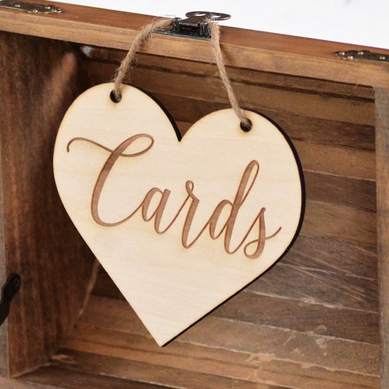 Puede incluir: Un letrero de madera en forma de coraz&oacute;n con la palabra "Cards" escrita en cursiva. El letrero est&aacute; colgado de una caja de madera con un cierre.