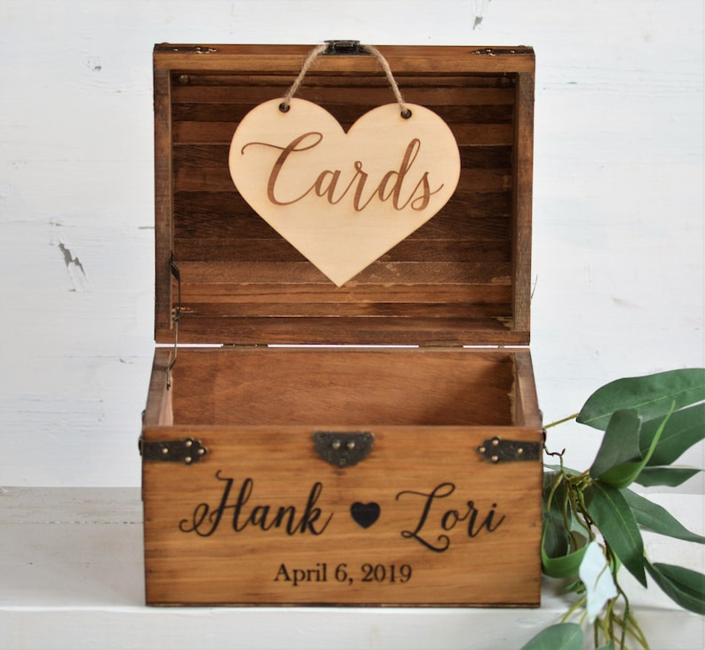 Puede incluir: Una caja de madera con un letrero en forma de coraz&oacute;n que dice "Cards" colgando de la parte superior. La caja est&aacute; decorada con detalles met&aacute;licos y tiene los nombres "Hank & Lori" y la fecha "April 6, 2019" grabados en la parte delantera.