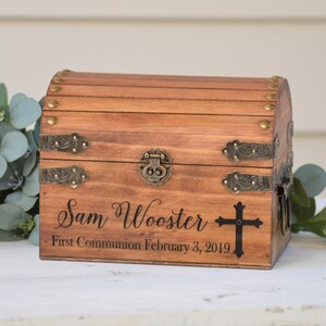 Puede incluir: Un cofre del tesoro de madera con detalles en lat&oacute;n y una inscripci&oacute;n personalizada que dice "Sam Wooster - Primera Comuni&oacute;n 3 de febrero de 2019" y una cruz.