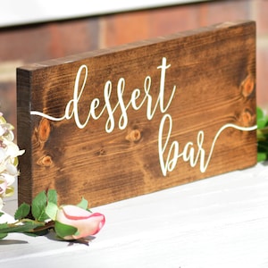 May include: A rustic wooden sign with the words "dessert bar" written in white cursive script. The sign is stained a dark brown color and has a natural wood grain.