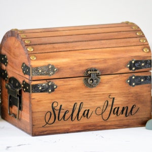 May include: A wooden treasure chest with brass accents and a hinged lid. The chest is personalized with the name "Stella Jane" in black lettering.
