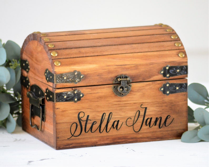 May include: A wooden treasure chest with brass accents and a hinged lid. The chest is personalized with the name "Stella Jane" in black lettering.