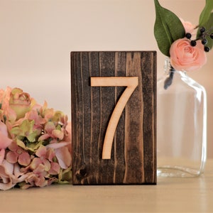 May include: A wooden table number with the number 7 displayed in a light wood color. The number is on a dark brown wooden block. The block is sitting on a white surface. There is a bouquet of pink flowers and a vase with a single pink flower to the left of the table number.
