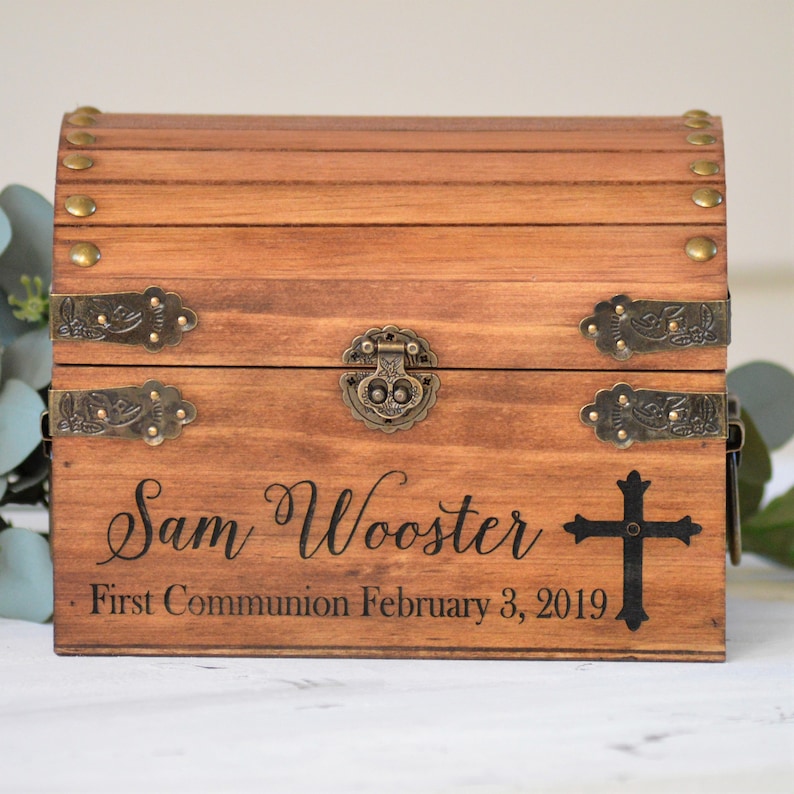 Puede incluir: Una caja de madera para recuerdos con tapa con bisagras y detalles en lat&oacute;n. La caja est&aacute; grabada con el nombre "Sam Wooster", el texto "Primera Comuni&oacute;n 3 de febrero de 2019" y una cruz.