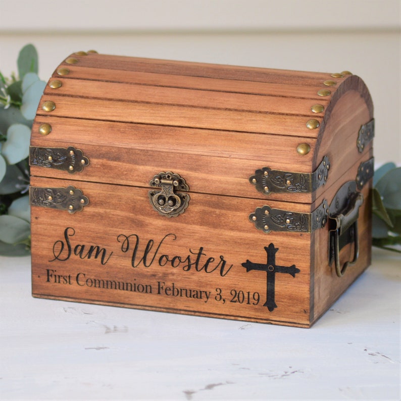 Puede incluir: Una caja de madera para recuerdos con tapa con bisagras y detalles en lat&oacute;n. La caja est&aacute; grabada con el nombre "Sam Wooster" y el texto "Primera Comuni&oacute;n 3 de febrero de 2019". Tambi&eacute;n hay una cruz negra grabada en la caja.