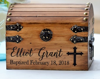 May include: A wooden chest with a curved lid, featuring brass-colored metal studs, hinges, and a central latch. The front is engraved with the name Elliot Grant, a cross symbol, and the text Baptized February 18, 2018, in a black script and serif font.