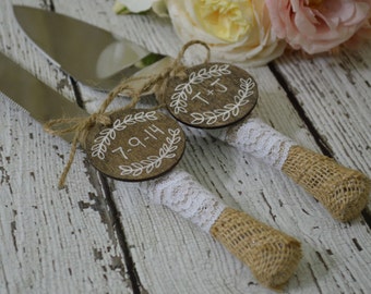 May include: Wedding cake serving set with stainless steel blades and burlap-wrapped handles. White lace and wooden tags with initials and date are attached. The tags have a white leaf design.