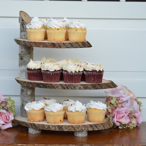 May include: A three-tiered wooden cupcake stand with a rustic, natural bark finish. The stand is decorated with colorful sprinkles and features cupcakes with white and red frosting.