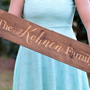 May include: A wooden sign with the text "The Kohlmen Family" engraved in a light brown color. The sign is held by a person wearing a light blue dress.