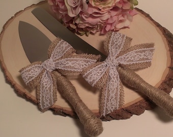 May include: A set of two cake cutting knives with burlap and lace bows. The knives are on a wooden slice.