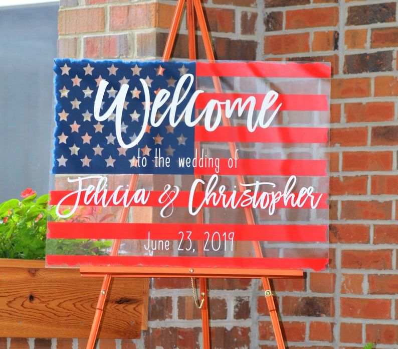 May include: A clear acrylic sign with a red, white and blue American flag design. The sign says "Welcome to the wedding of Felicia & Christopher June 23, 2019".