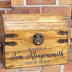 May include: A wooden keepsake box with brass hinges and a lock. The box is engraved with the text "In memory of Tom Klingensmith 12/27/58 - 09/18/21".