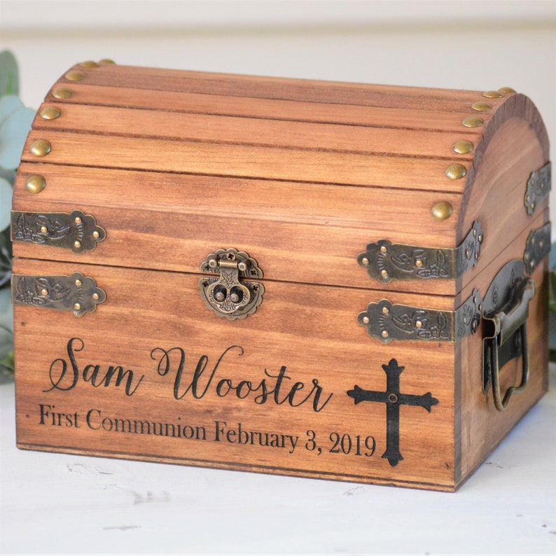 Puede incluir: Una caja de madera para recuerdos con tapa con bisagras y detalles met&aacute;licos. La caja est&aacute; personalizada con el nombre "Sam Wooster" y el texto "Primera Comuni&oacute;n 3 de febrero de 2019". Una cruz negra est&aacute; grabada en la parte delantera de la caja.