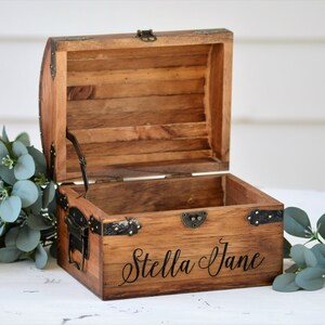 May include: A wooden treasure chest with a hinged lid and metal accents. The chest is open and the inside is empty. The chest is engraved with the name "Stella Jane".