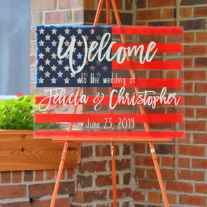 May include: Welcome to the wedding of Felicia & Christopher, June 23, 2019. A clear acrylic sign with a red, white and blue American flag design.