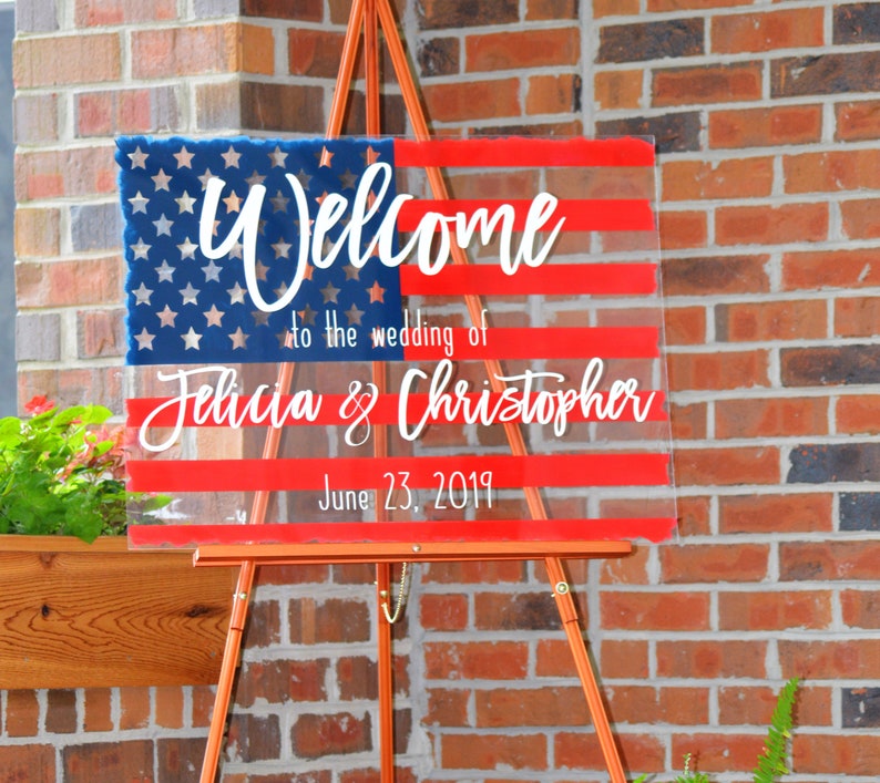 May include: Welcome to the wedding of Felicia & Christopher, June 23, 2019. A clear acrylic sign with a red, white and blue American flag design.