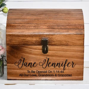 May include: A medium-brown wooden chest with a curved lid and a brass-colored metal latch. The front features black text reading: Jane Jennifer, To Be Opened On 1-14-44, All Our Love, Grandmom & Granddad. The box has a visible wood grain texture.