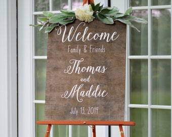May include: A wooden welcome sign with white lettering on a brown easel. The sign reads "Welcome Family & Friends Thomas and Maddie July 13, 2019".