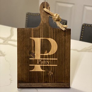 K&ouml;nnte beinhalten: Ein dunkelbraunes Holzbrett zum Schneiden mit einem gro&szlig;en wei&szlig;en Buchstaben "P" und dem Namen "Patty" eingraviert. Das Brett hat einen Griff mit einem geknoteten Seil.